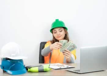 Mulher com capacete verde e colete laranja conta cédulas de dinheiro sentada a uma mesa com notebook e capacetes de obra