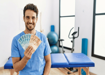 Profissional de saúde em scrub azul segura notas de 100 e 50 reais em clínica com maca e bicicleta ergométrica, simbolizando as opções renda extra para fisioterapeutas.