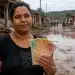 Mulher atingida por enchente segura notas de dinheiro em área destruída, representando os próximos repasses do Auxílio Reconstrução.