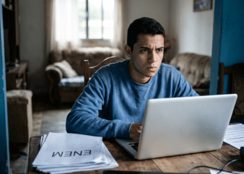 Jovem usa um notebook concentrado à mesa e, ao redor, papéis com a palavra Enem e a sala de uma casa