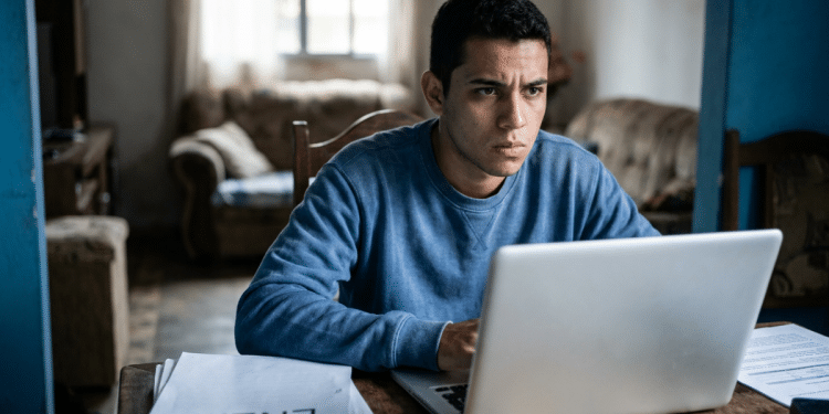 Jovem usa um notebook concentrado à mesa e, ao redor, papéis com a palavra Enem e a sala de uma casa