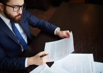 Homem de terno azul e óculos examina documentos sobre mesa escura segurando caneta e folha, simbolizando o salário de um perito avaliador judicial.