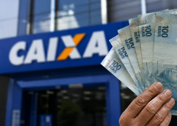 Mão segurando cédulas de cem reais em frente a uma agência da Caixa, em referência ao Saque Calamidade do FGTS de até R$ 6.220 liberado para novos municípios em 2026.
