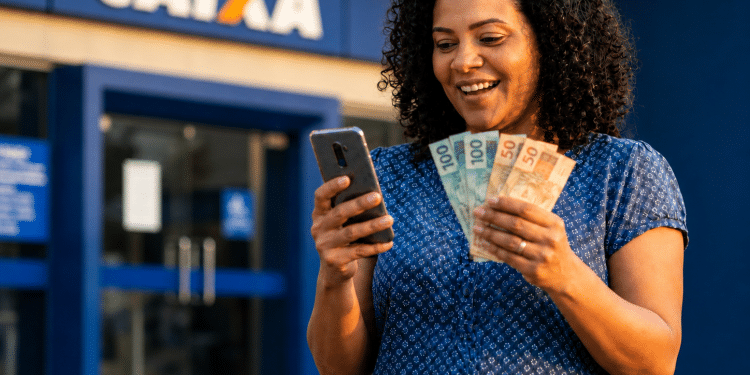 Mulher sorri em frente a uma agência da Caixa ao solicitar pelo celular o Saque Calamidade do FGTS e segura notas de R$ 100 e R$ 50