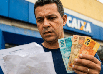 Homem confere papel em frente a uma agência da Caixa segurando notas de R$ 100, R$ 50 e R$ 20, em referência ao Saque Calamidade do FGTS.