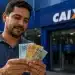 Homem conferindo cédulas de real em frente à Caixa, representando saque de até R$ 6.220 liberado para solicitação nesta terça-feira, 19 de maio.