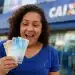 Mulher feliz segurando notas de dinheiro em frente a uma agência da CAIXA, destacando saque de R$ 6.220 disponível até 30 de maio.