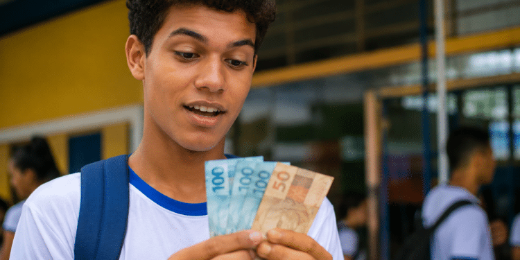 Estudante de uniforme conta cédulas de real em frente a uma escola