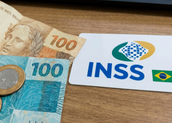 Cartão branco com logo INSS e bandeira do Brasil ao lado de notas de 100 reais e moedas sobre superfície de madeira, simbolizando a segunda parcela do 13º salário para quem recebe até R$ 1.621, atual salário mínimo.
