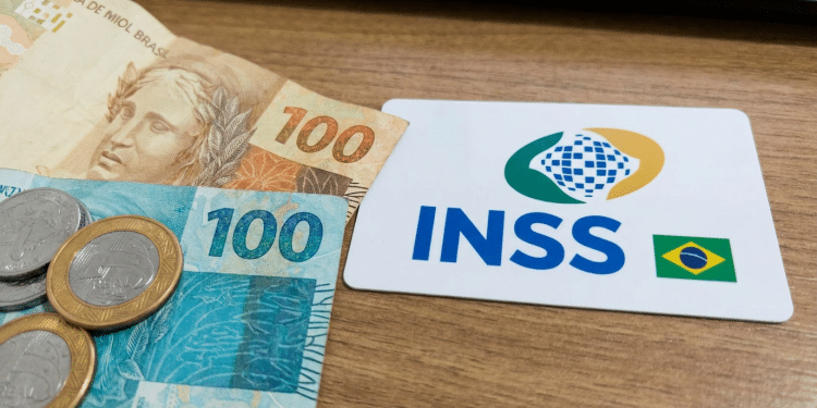 Cartão branco com logo INSS e bandeira do Brasil ao lado de notas de 100 reais e moedas sobre superfície de madeira, simbolizando a segunda parcela do 13º salário para quem recebe até R$ 1.621, atual salário mínimo.