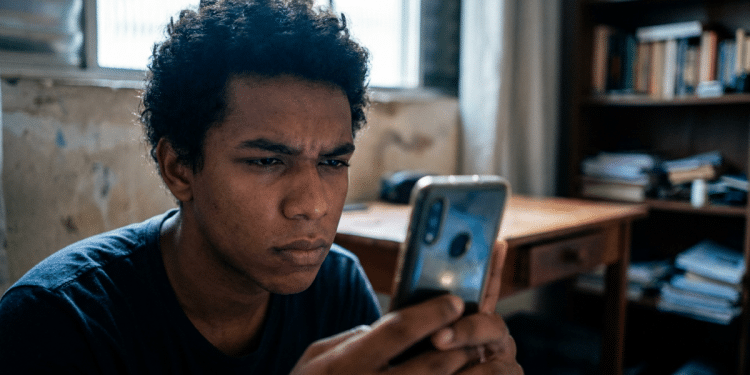 Jovem usa o celular com expressão concentrada e, ao redor, a escrivaninha e a estante de um quarto