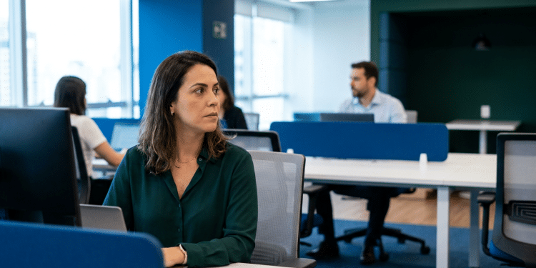 Mulher de blusa verde observa colegas ao fundo em escritório de espaço compartilhado.