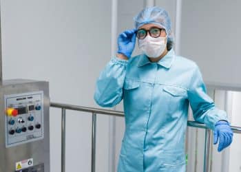 Profissional com jaleco azul, touca, máscara e luvas ajusta óculos próximo a painel de controle em sala limpa industrial, simbolizando a profissão de