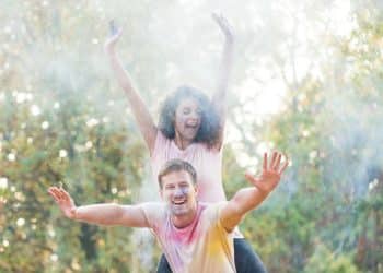 Casal sorri de braços erguidos ao ar livre, cercado por pó colorido suspenso no ar.