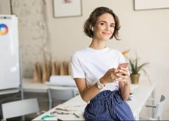Mulher jovem sorrindo segura o celular em ambiente de trabalho
