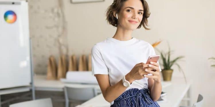 Mulher jovem sorrindo segura o celular em ambiente de trabalho