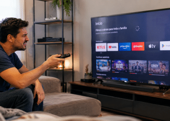 Homem de camiseta azul sentado no sofá aponta controle remoto para Smart TV com apps de streaming
