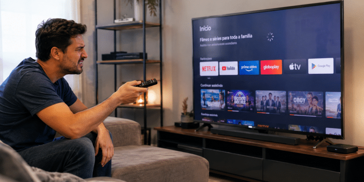 Homem de camiseta azul sentado no sofá aponta controle remoto para Smart TV com apps de streaming