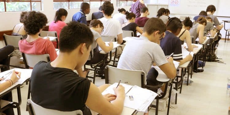 Sala de aula com diversos estudantes sentados em carteiras realizando uma prova com folhas e canetas