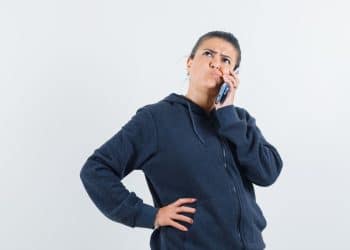 Mulher pensativa com expressão curiosa ao telefone em fundo claro.