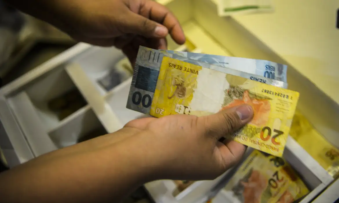 Mãos seguram notas de 100 e 20 reais sobre caixa registradora aberta com cédulas, simbolizando o pagamento do BPC/INSS.