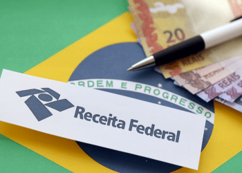 Papel com logo e nome Receita Federal sobre bandeira do Brasil, notas de real e caneta.