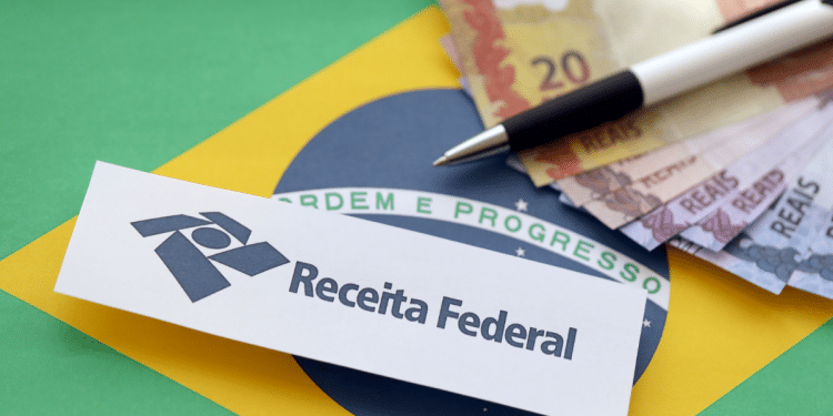 Papel com logo e nome Receita Federal sobre bandeira do Brasil, notas de real e caneta.