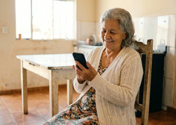 Idosa sorridente usa celular em casa simples.