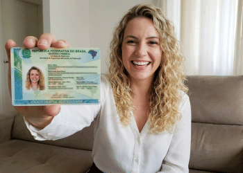 Mulher loira com cabelos cacheados mostra Carteira de Identidade Nacional do Brasil sentada em sofá.