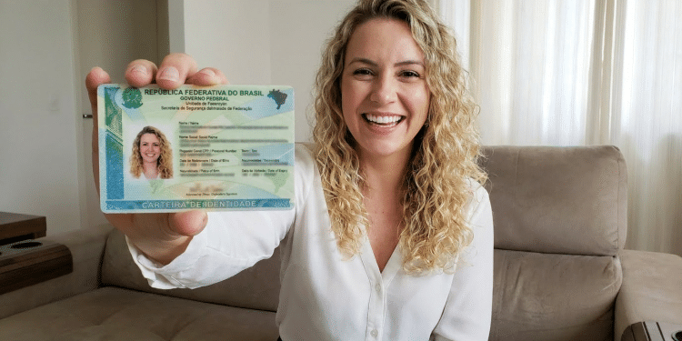 Mulher loira com cabelos cacheados mostra Carteira de Identidade Nacional do Brasil sentada em sofá.