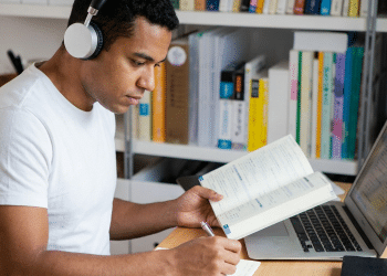 Homem jovem com fone de ouvido estudando com livro aberto e notebook em mesa de madeira.