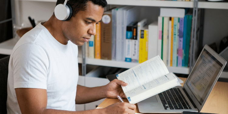 Homem jovem com fone de ouvido estudando com livro aberto e notebook em mesa de madeira.