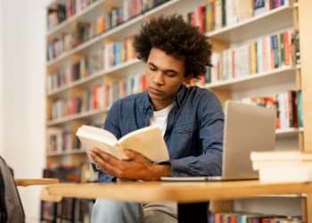 Jovem lendo livro em biblioteca rodeado por tecnologia e livros.
