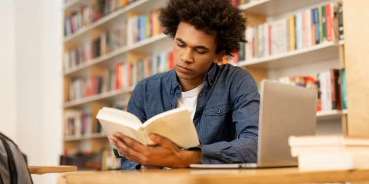Jovem lendo livro em biblioteca rodeado por tecnologia e livros.