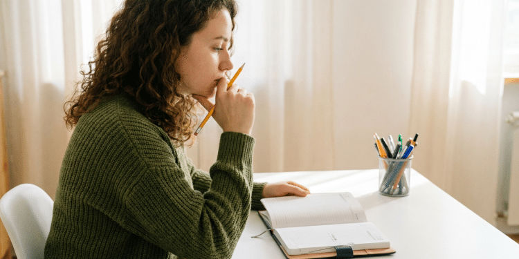 Jovem pensativa estuda com caderno e lápis em ambiente caseiro.