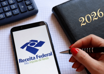 Celular com app Receita Federal, calculadora cinza, agenda preta 2026 e mão feminina com caneta sobre papel.
