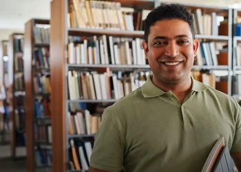 Homem segurando livros em biblioteca, representando estudante que pode ingressar em curso técnico com certificação do Encceja.