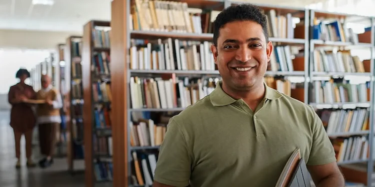 Homem segurando livros em biblioteca, representando estudante que pode ingressar em curso técnico com certificação do Encceja.