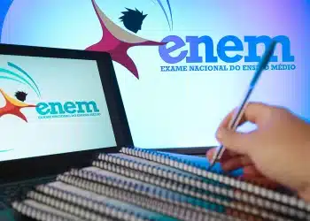 Logo do Enem em tela de notebook e monitor, mão segurando caneta sobre cadernos espiralados, simbolizando a utilização das notas do Enem.