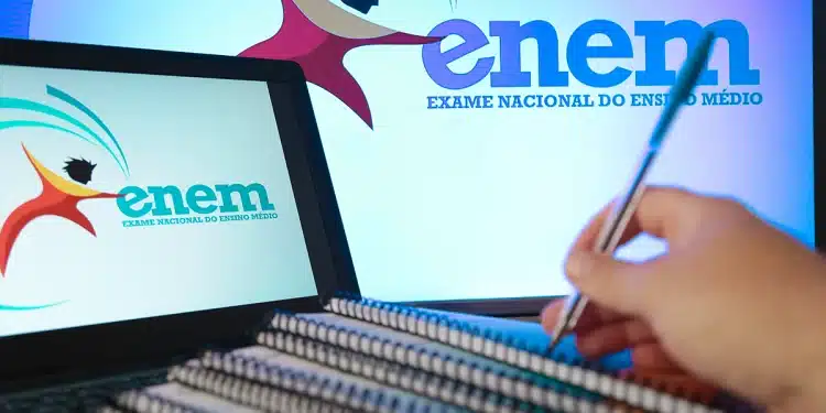 Logo do Enem em tela de notebook e monitor, mão segurando caneta sobre cadernos espiralados, simbolizando a utilização das notas do Enem.
