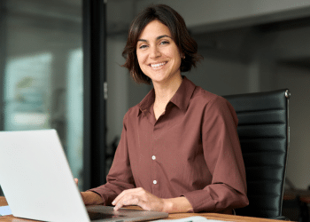 Mulher sorridente trabalha em escritório moderno diante de notebook.