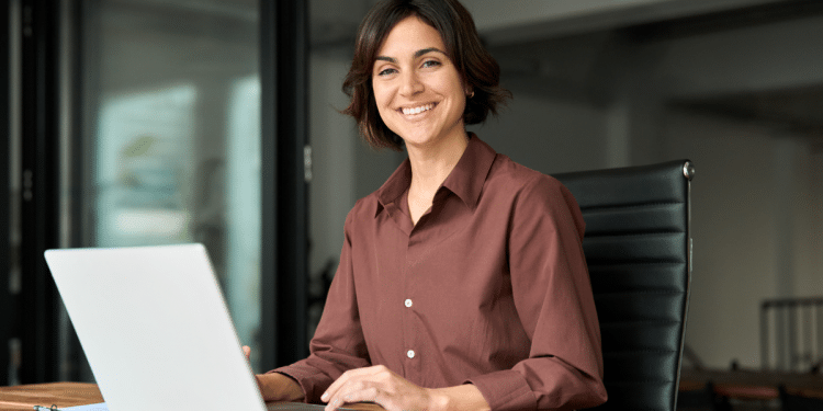 Mulher sorridente trabalha em escritório moderno diante de notebook.