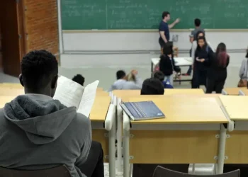 Estudante em sala de aula universitária acompanhando conteúdo em quadro verde durante aula.