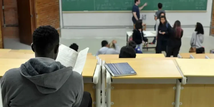 Estudante em sala de aula universitária acompanhando conteúdo em quadro verde durante aula.