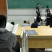 Estudante em sala de aula universitária acompanhando conteúdo em quadro verde durante aula.