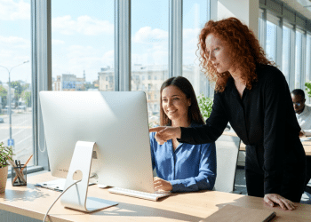 Duas mulheres trabalhando juntas em frente a computador, uma aponta para tela, escritório iluminado com janelas amplas.