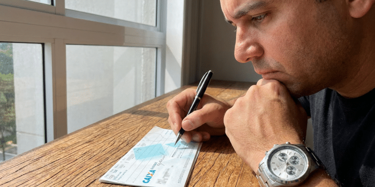 Homem pensativo preenche cheque da Caixa Econômica Federal.
