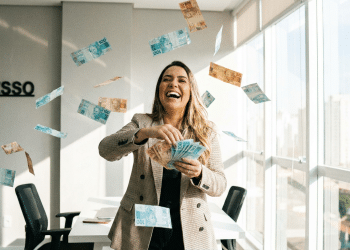 Mulher sorridente comemora conquista jogando dinheiro em escritório com placa "SUCESSO".