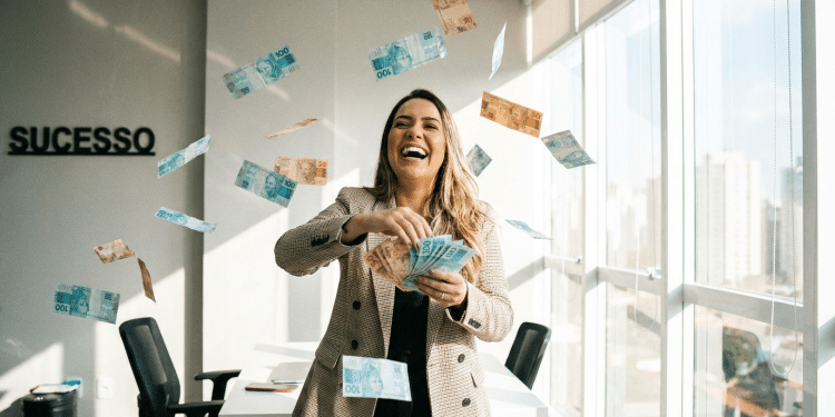 Mulher sorridente comemora conquista jogando dinheiro em escritório com placa "SUCESSO".