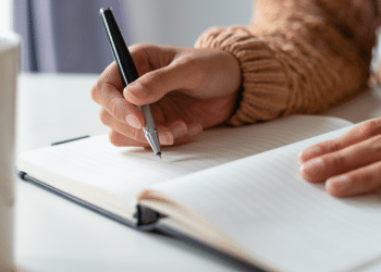 Pessoa escrevendo em caderno de folhas pautadas em ambiente interno.